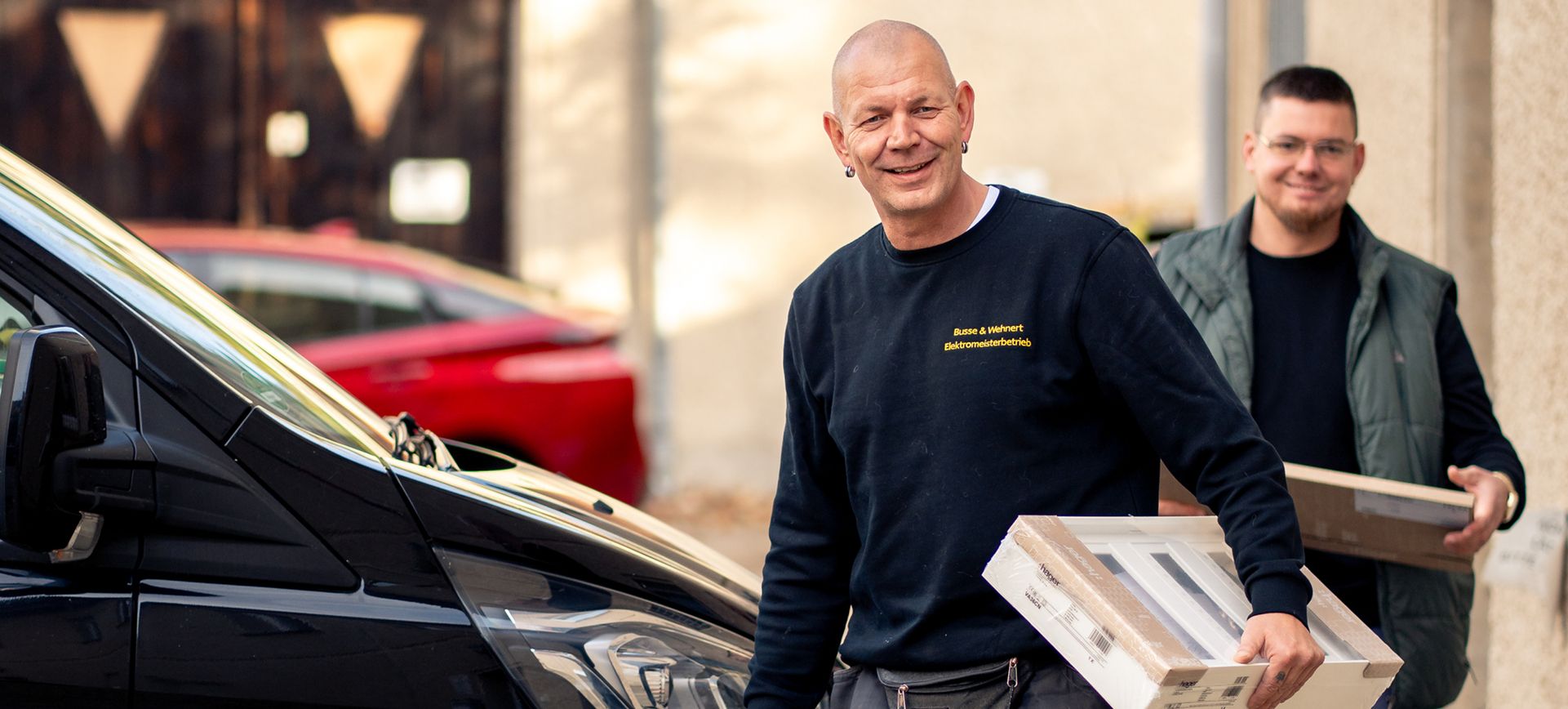 Zwei Männer tragen Pakete neben einem schwarzen Fahrzeug auf einer Straße mit parkenden Autos im Hintergrund